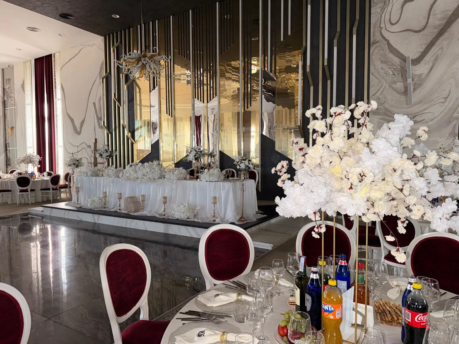 Prezidiu de nuntă în alb, cu aranjamente florale, într-o sală modernă cu pereți oglindă
