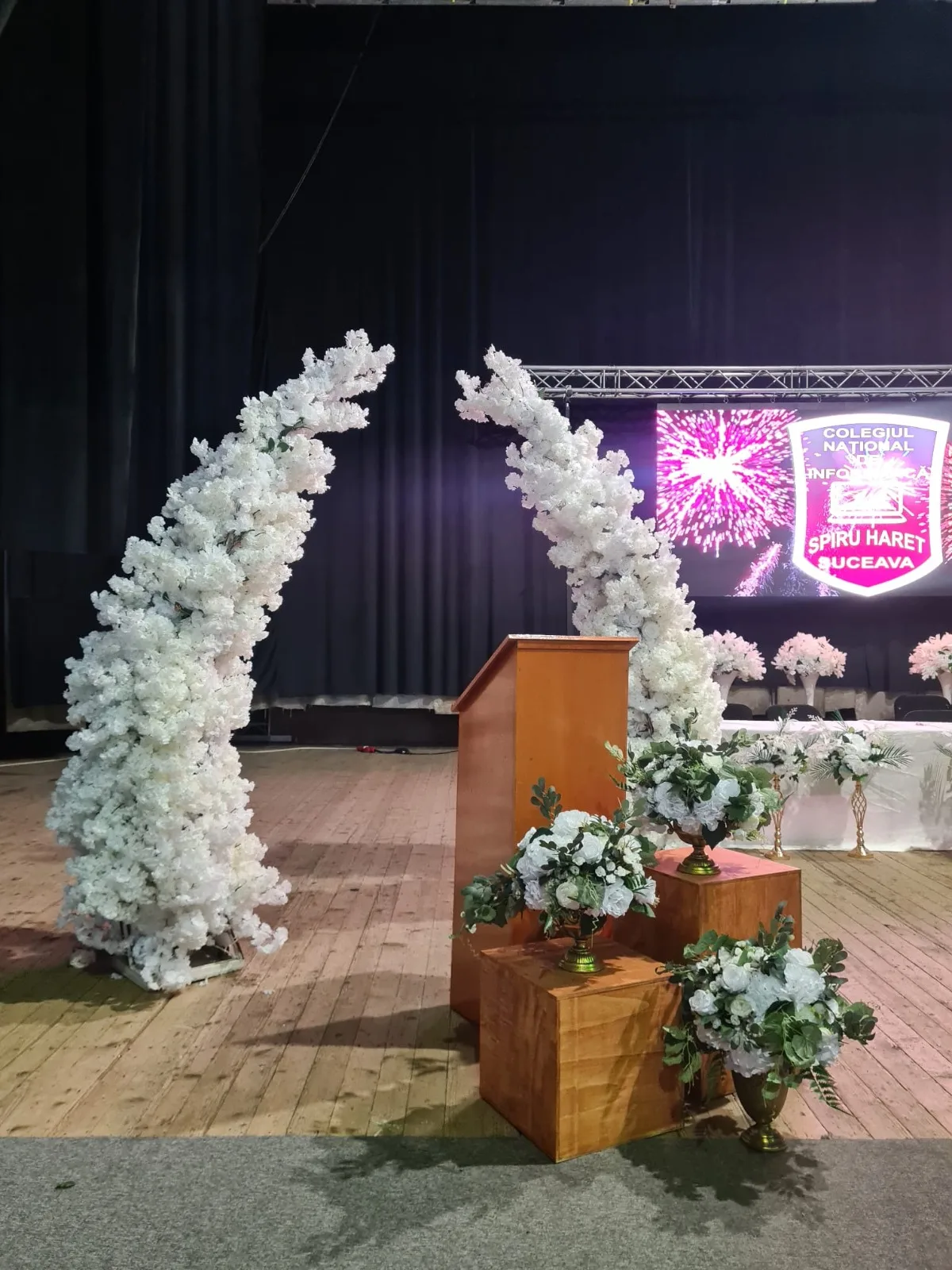 Arcade de flori albe langa podium pe scena, decor cu artificii pe ecran in fundal