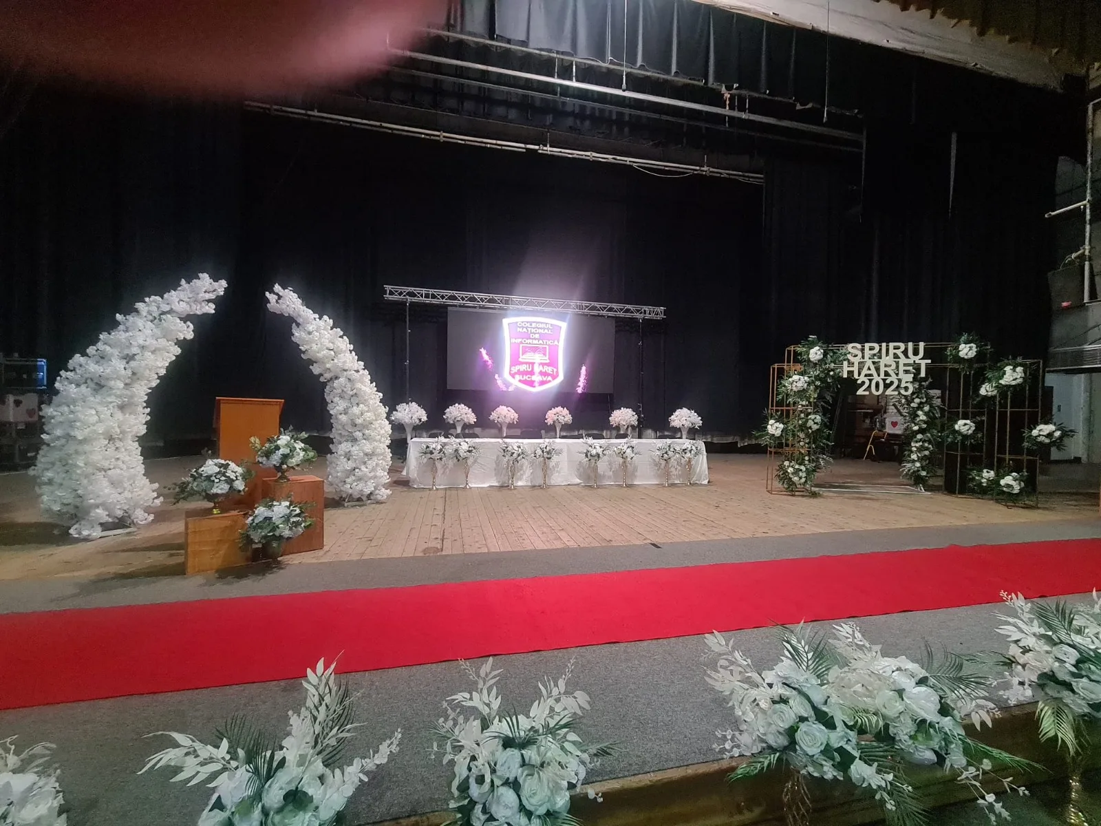 Arcade de flori albe langa podium din lemn pe scena, banner Spiru Haret 2025 pe ecran