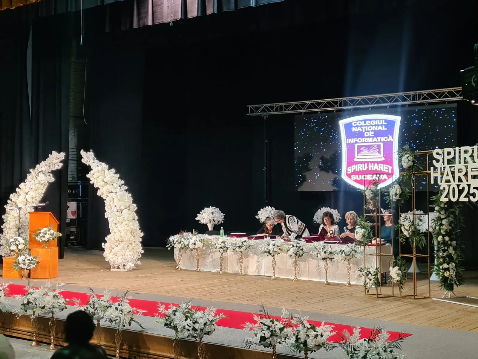 Scena cu prezidiu decorat cu arcade de flori albe si verdeata, banner Spiru Haret 2025