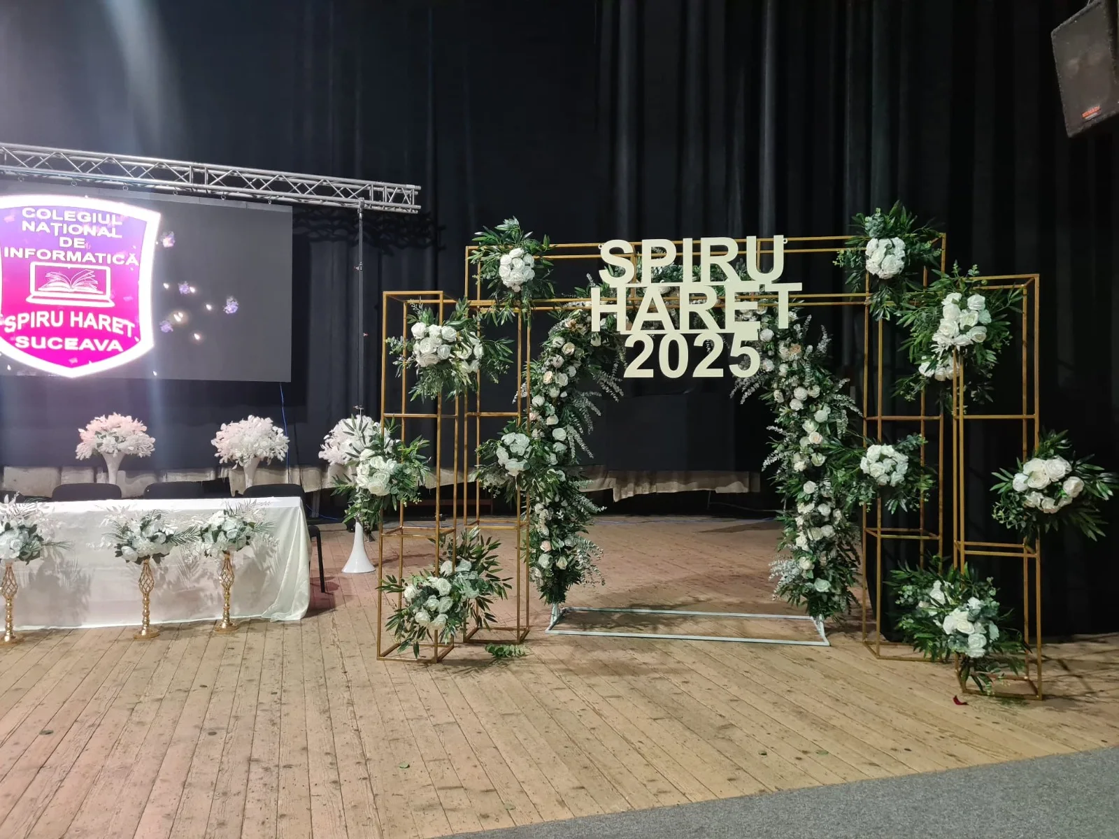 Structuri metalice aurii cu flori albe si verdeata pe scena, banner Spiru Haret 2025