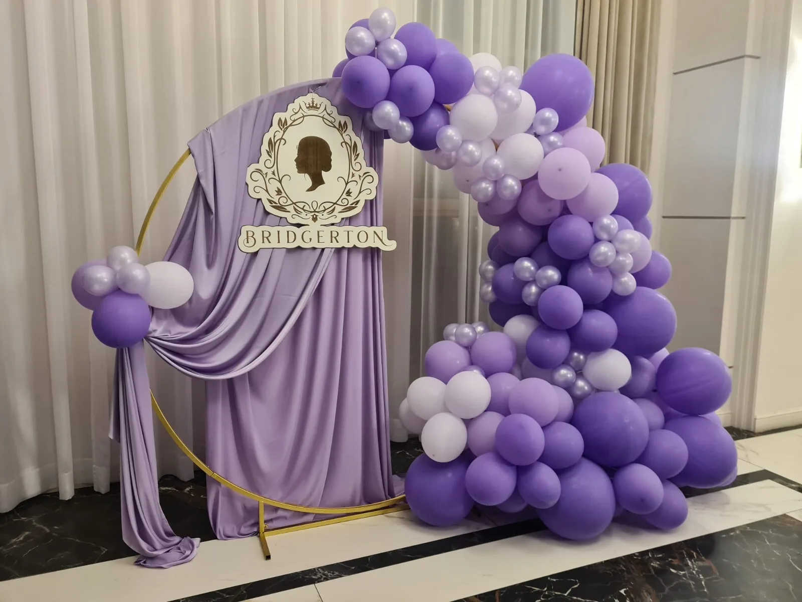 Decor tematic Bridgerton cu draperie mov, blazon auriu si arcada mare de baloane mov si albe in interior