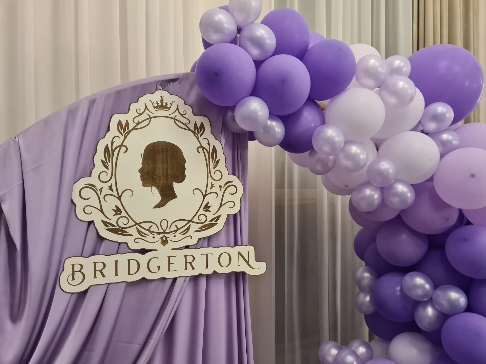 Decor tematic Bridgerton cu draperie mov, blazon auriu si arcada cu baloane mov si albe