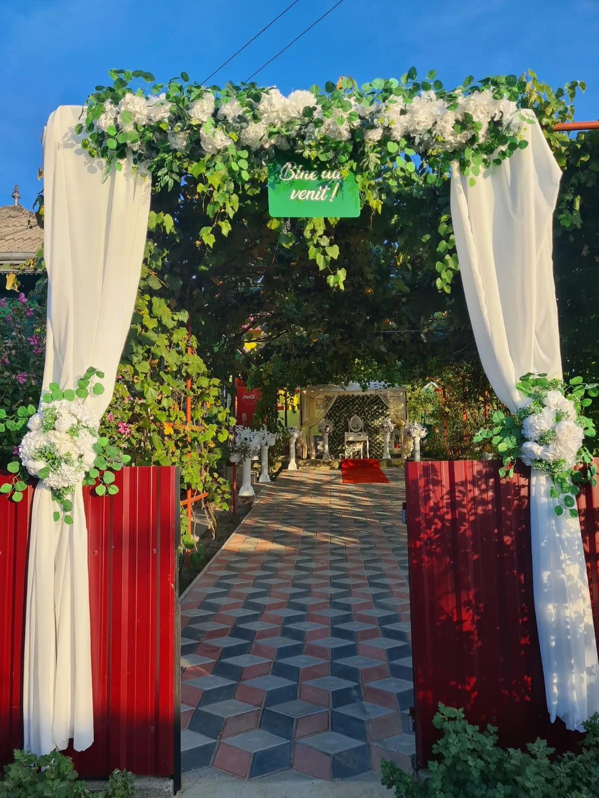 Arcadă florală alb-verde cu voaluri albe la poarta roșie, decor acasă la mireasă