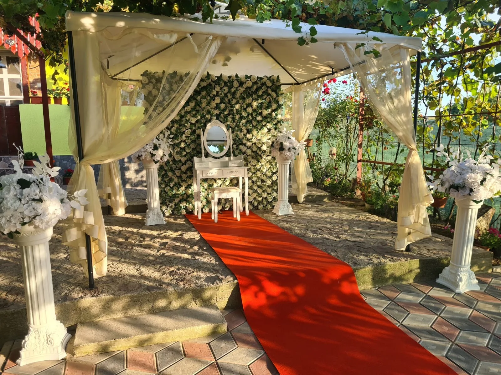 Colț foto cu perete floral alb-verde, măsuță cu oglindă și covor roșu, decor acasă la mireasă