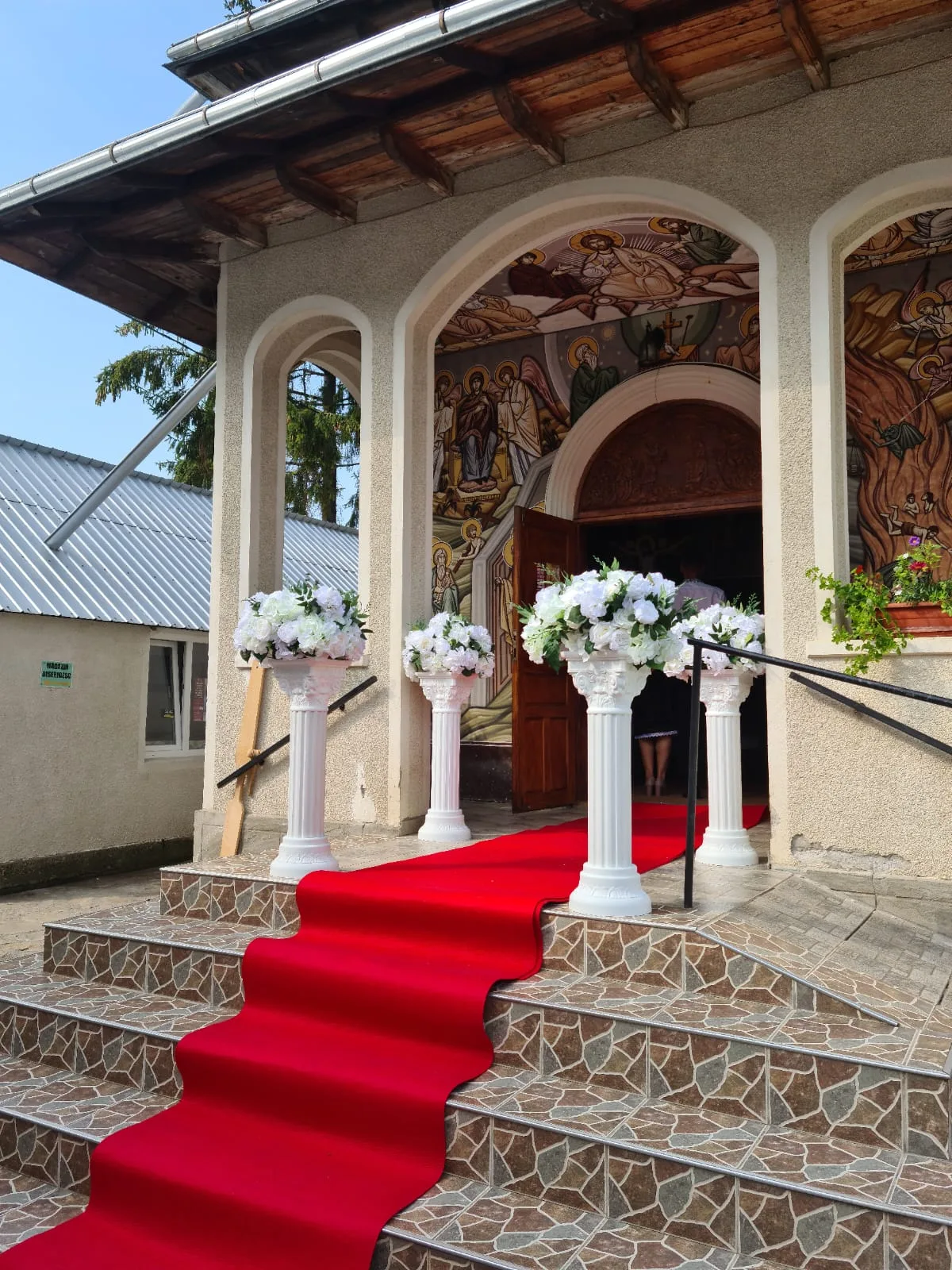 Scări cu covor roșu și coloane cu aranjamente florale albe la intrarea bisericii