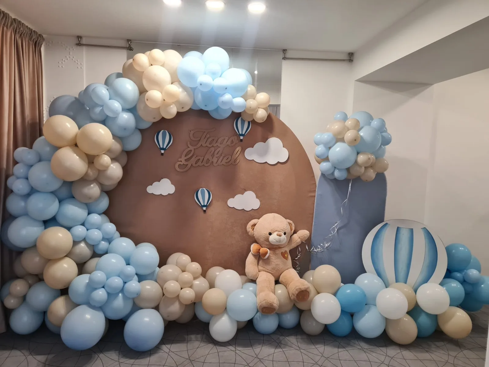 Photocorner de botez cu nori și montgolfiere, baloane bleu și crem, ursuleț de pluș, panoul cu numele Tiago Gabriel
