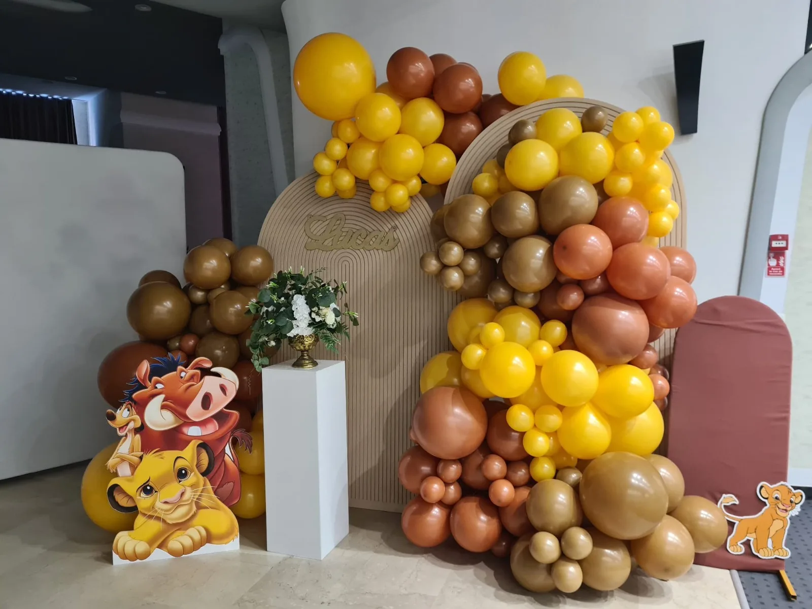Arcadă de baloane galbene și maro cu panoul numelui Lucas, siluete Timon și Pumba din Regele Leu, aranjament floral alb