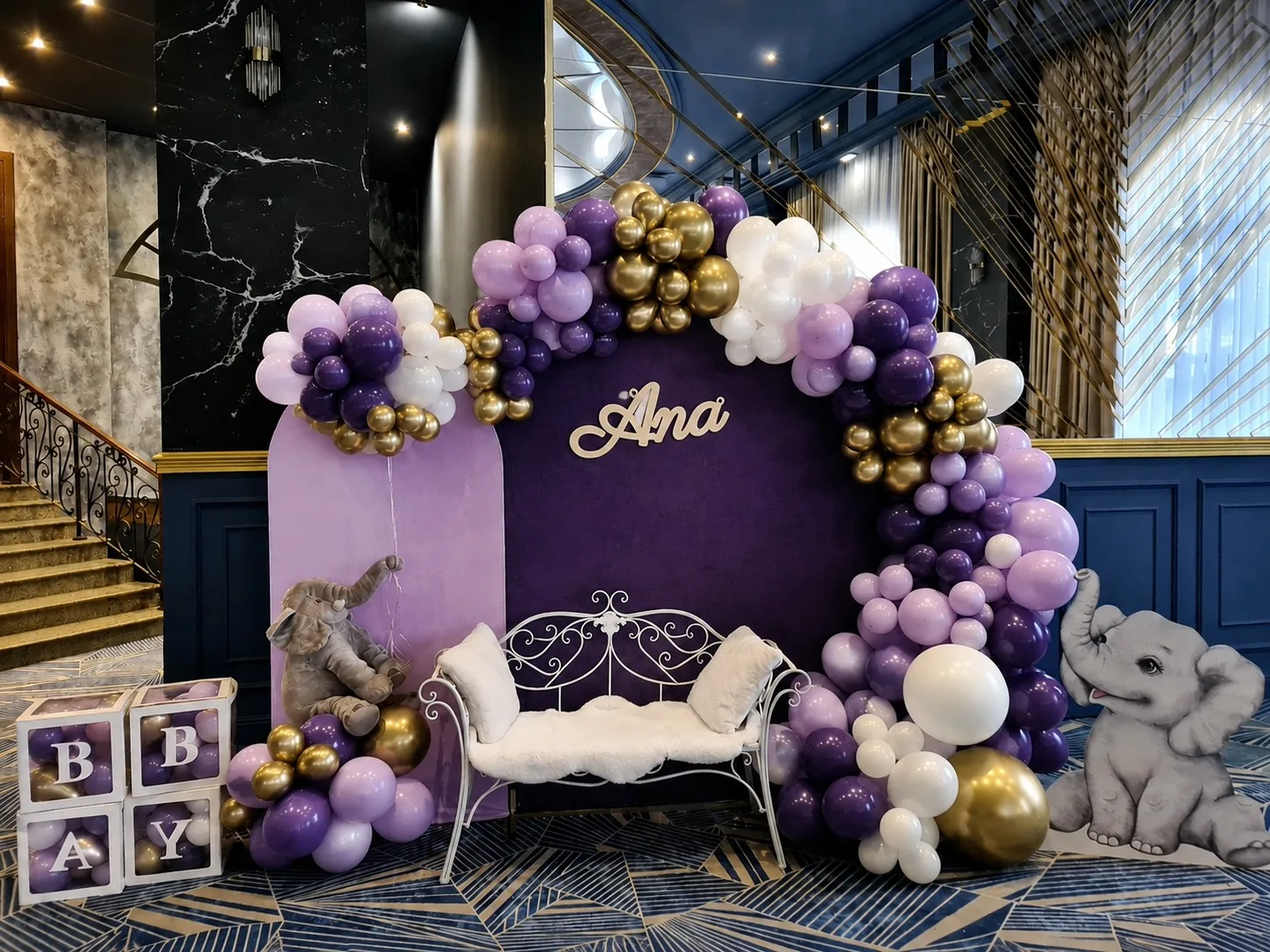 Arcadă de baloane mov, albe și aurii cu elefănței decorativi și bancă albă, panoul cu numele Ana