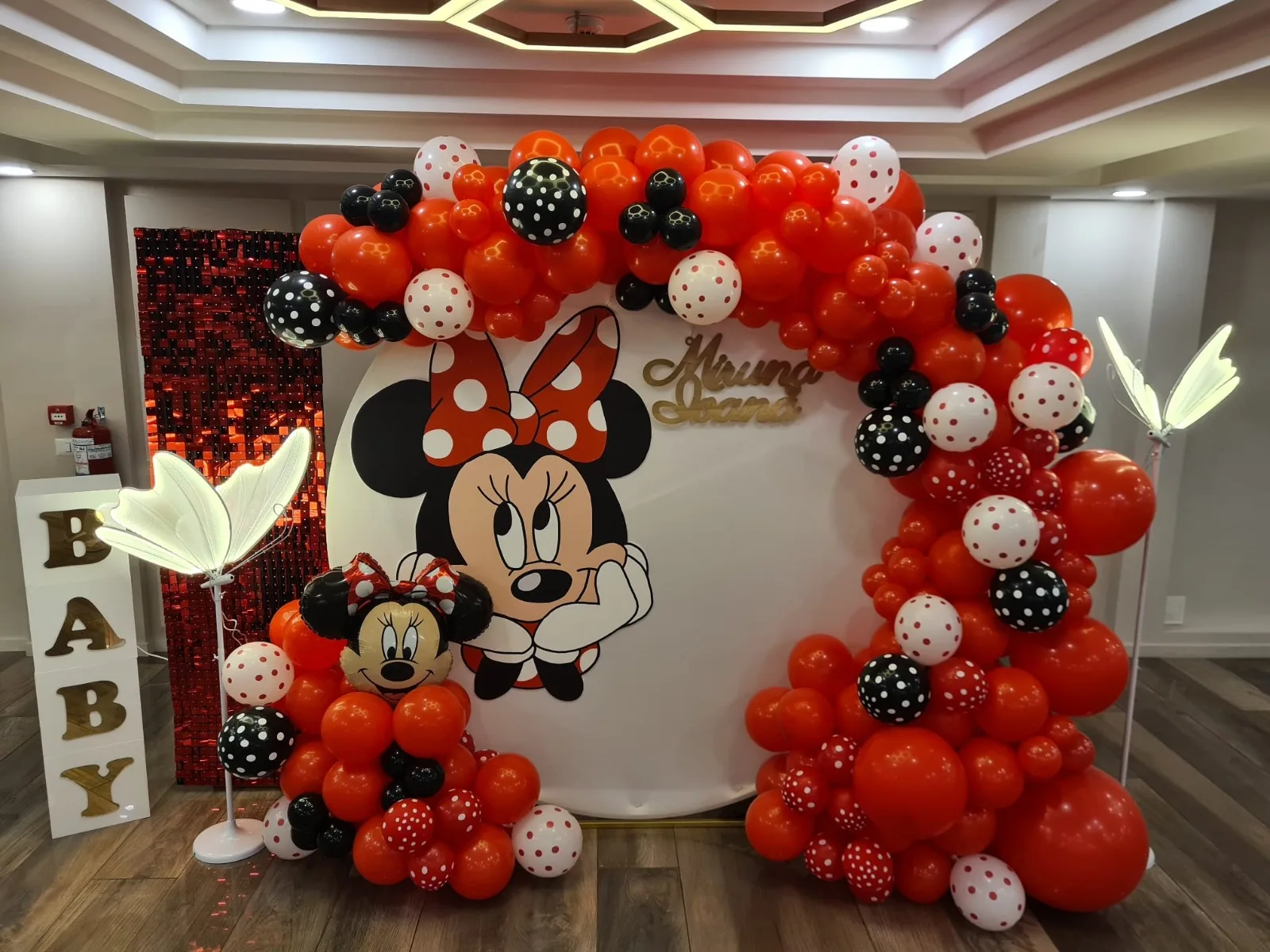Arcadă de baloane roșii, albe și negre cu buline, panou rotund cu Minnie Mouse și numele Miruna Ioana, fluturi luminoși