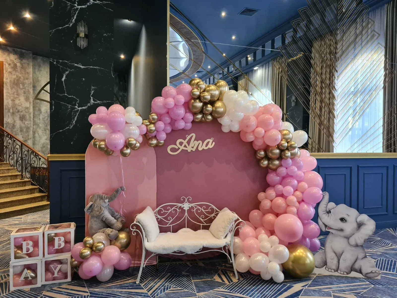 Arcadă de baloane roz și aurii cu elefănței decorativi și bancă albă, panoul cu numele Ana