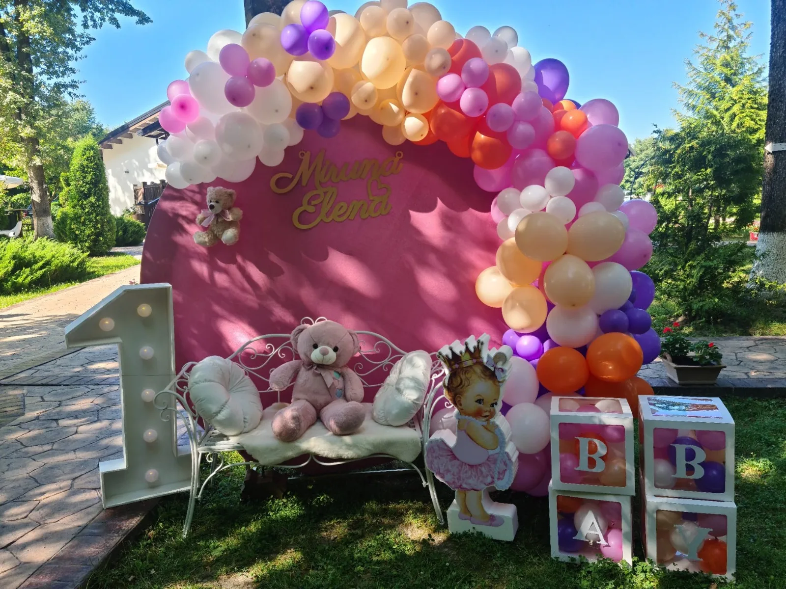 Backdrop roz într-o grădină, cu arcadă de baloane multicolore, bancă albă și numele Miruna Elena