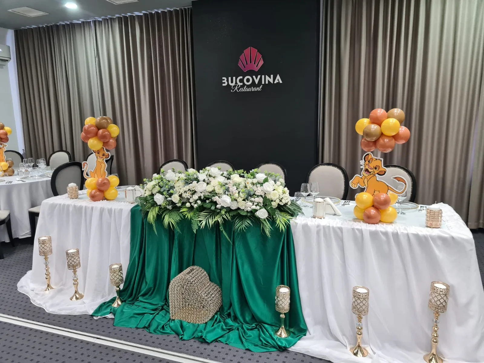 Masă festivă tematică Regele Leu, cu baloane maro, galbene și portocalii și siluete Simba, flori albe și restaurant Bucovina în fundal