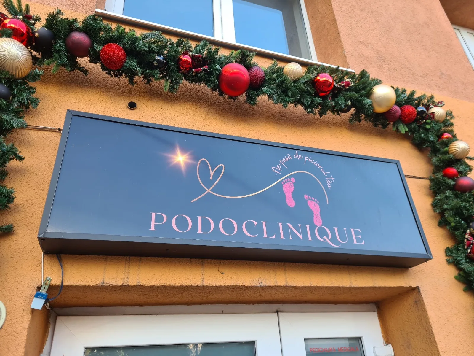 Ghirlanda de Craciun cu globuri rosii si aurii deasupra firmei Podoclinique