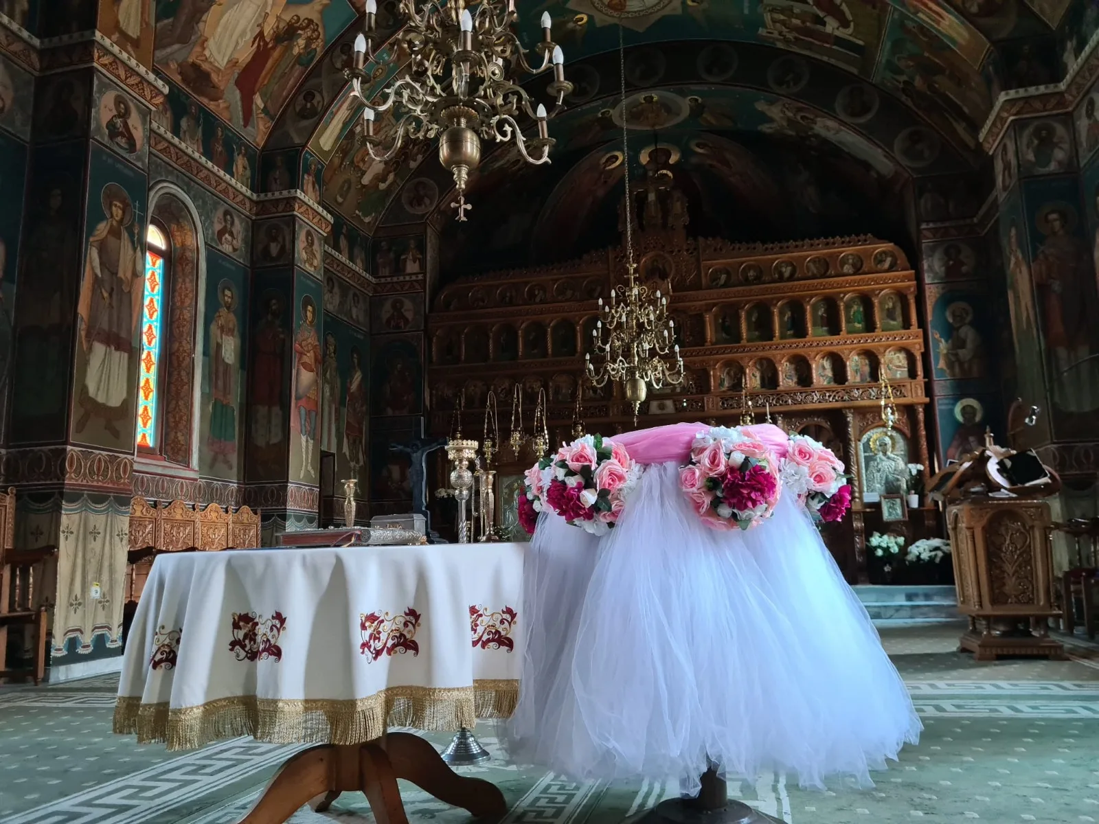 Decor de cristelniță în biserică, cu tul alb și coroniță de flori roz, sub candelabru