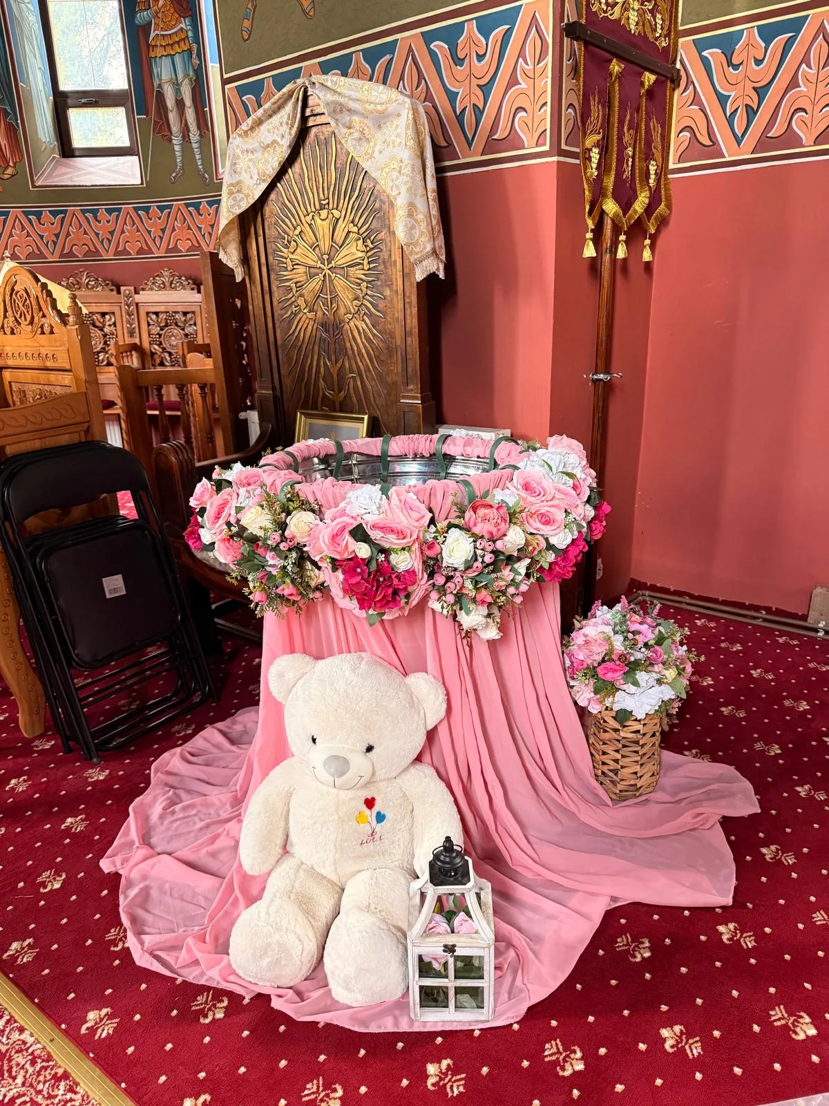 Cristelniță cu tul roz și coroană de flori, lângă iconostas, cu ursuleț de pluș și coș floral