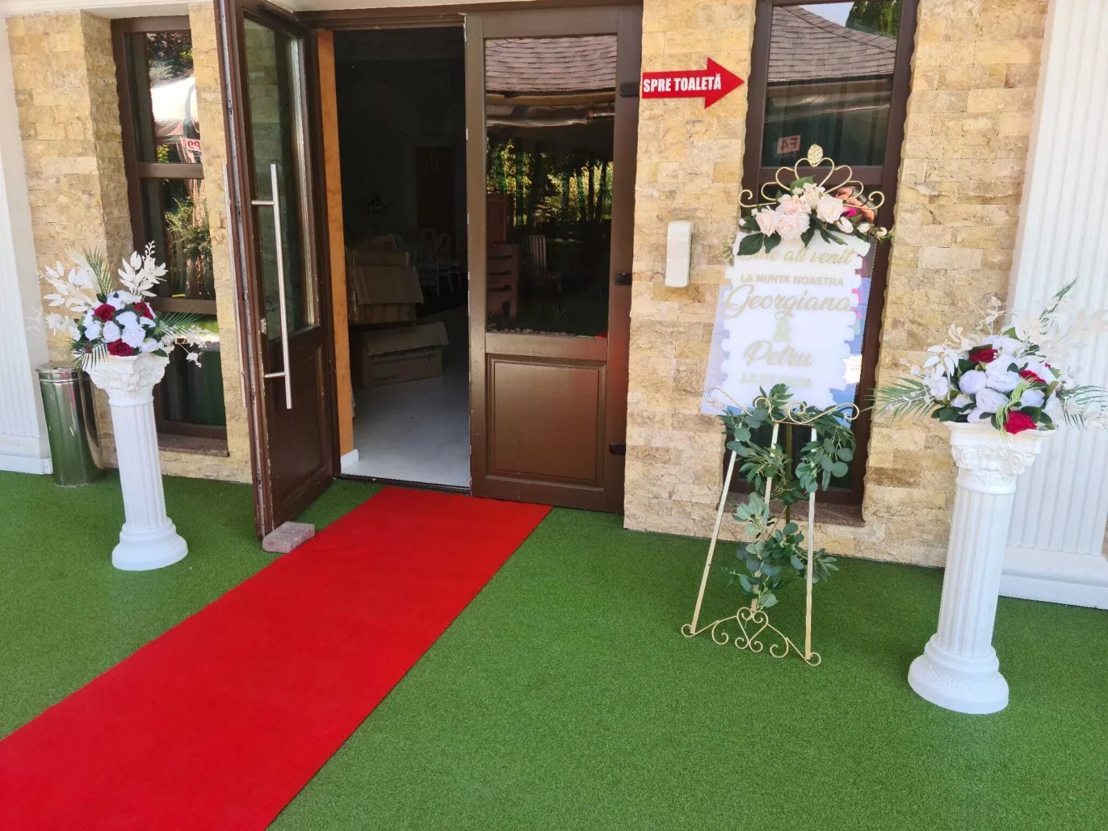 Decor intrare nuntă cu covor roșu, coloane albe cu aranjamente florale și panou cu mesaj de bun venit