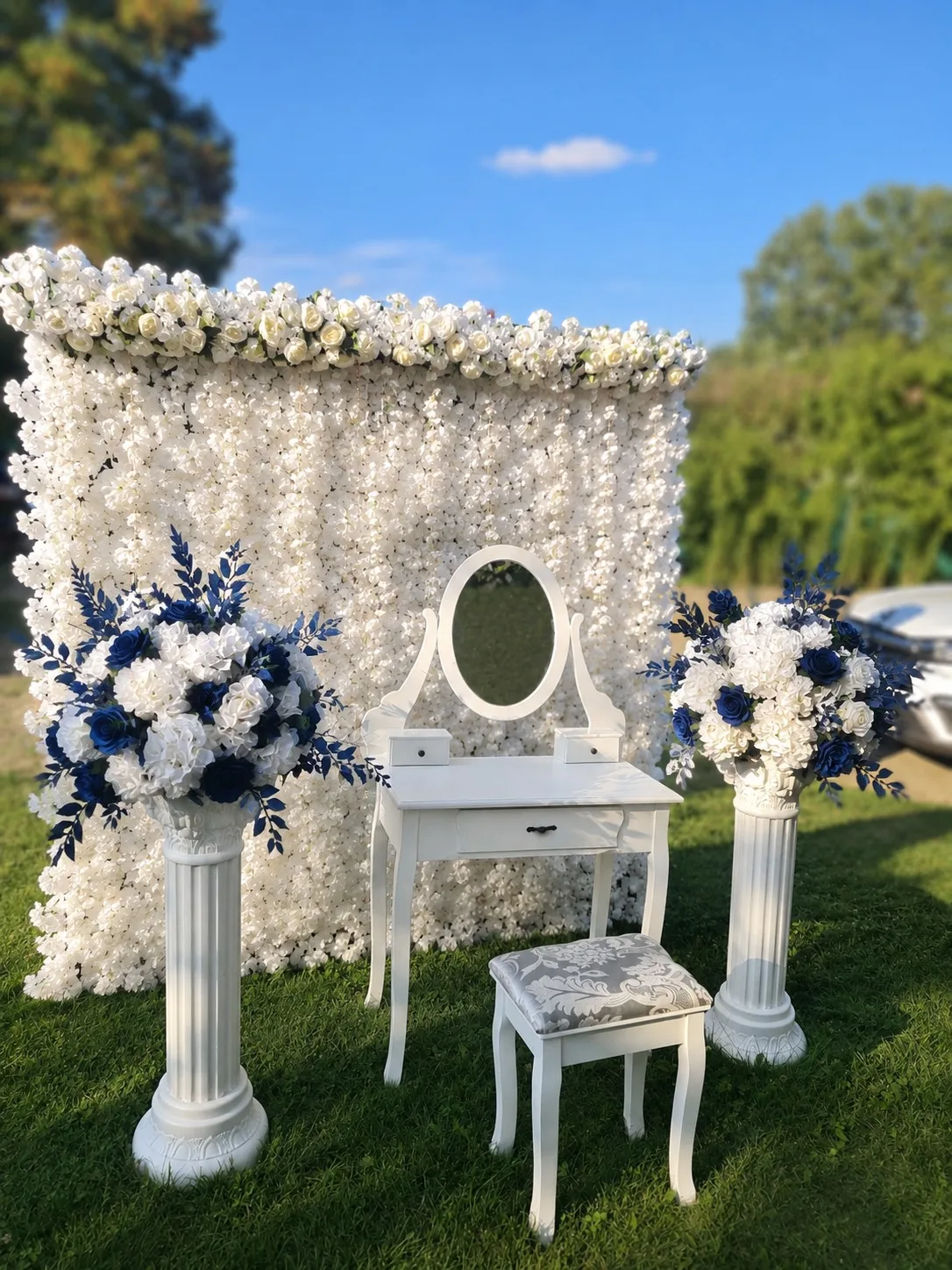 Măsuță de machiaj albă cu oglindă ovală, scăunel și coloane cu flori albe și albastru închis, în fața unui perete floral alb