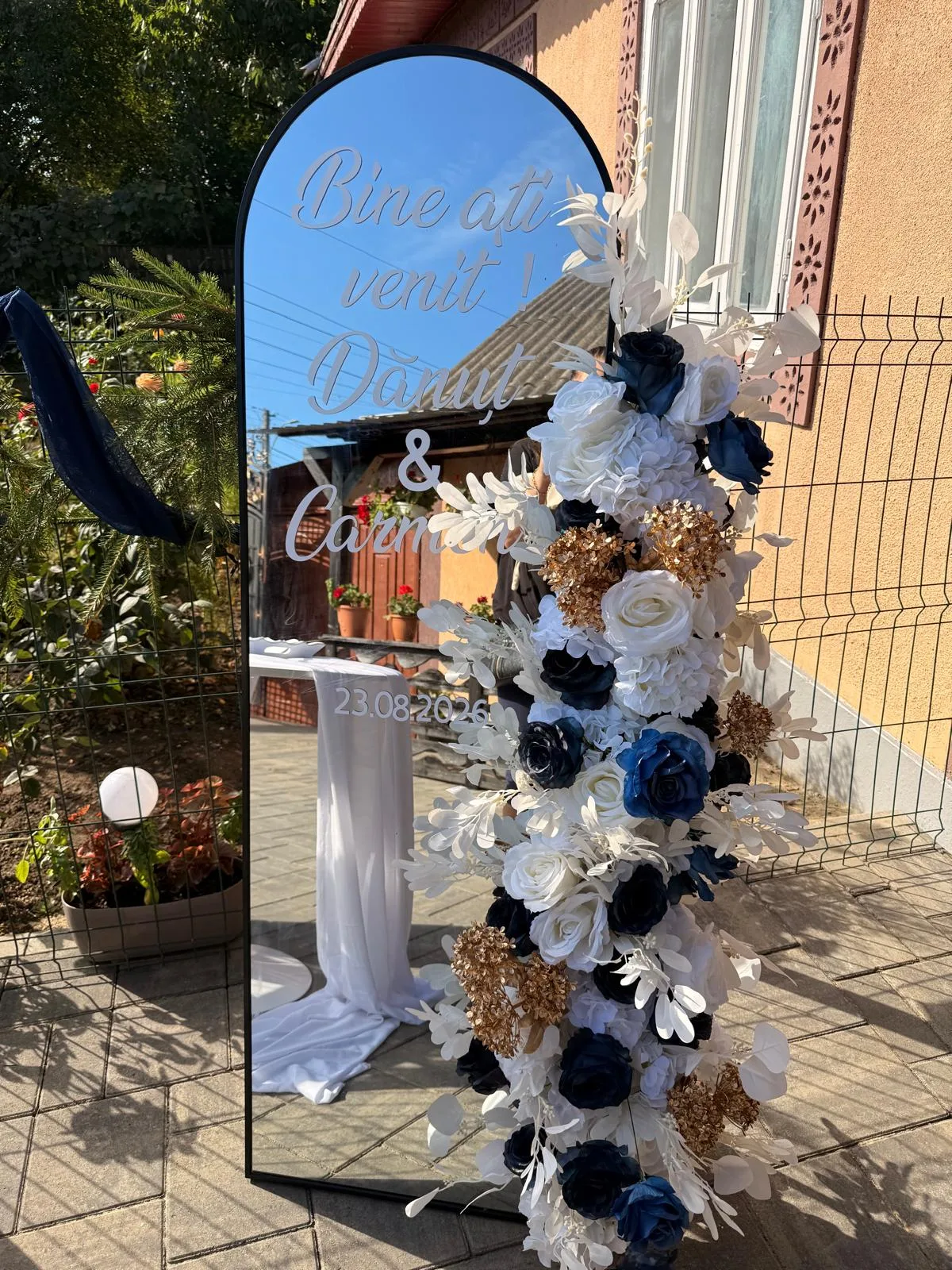 Măsuță albă rotundă lângă oglinda personalizată cu numele mirilor, decor pentru invitați