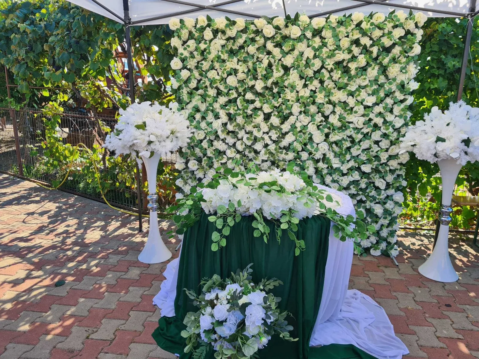 Perete floral alb cu verdeață, masă cu material verde și aranjamente florale albe