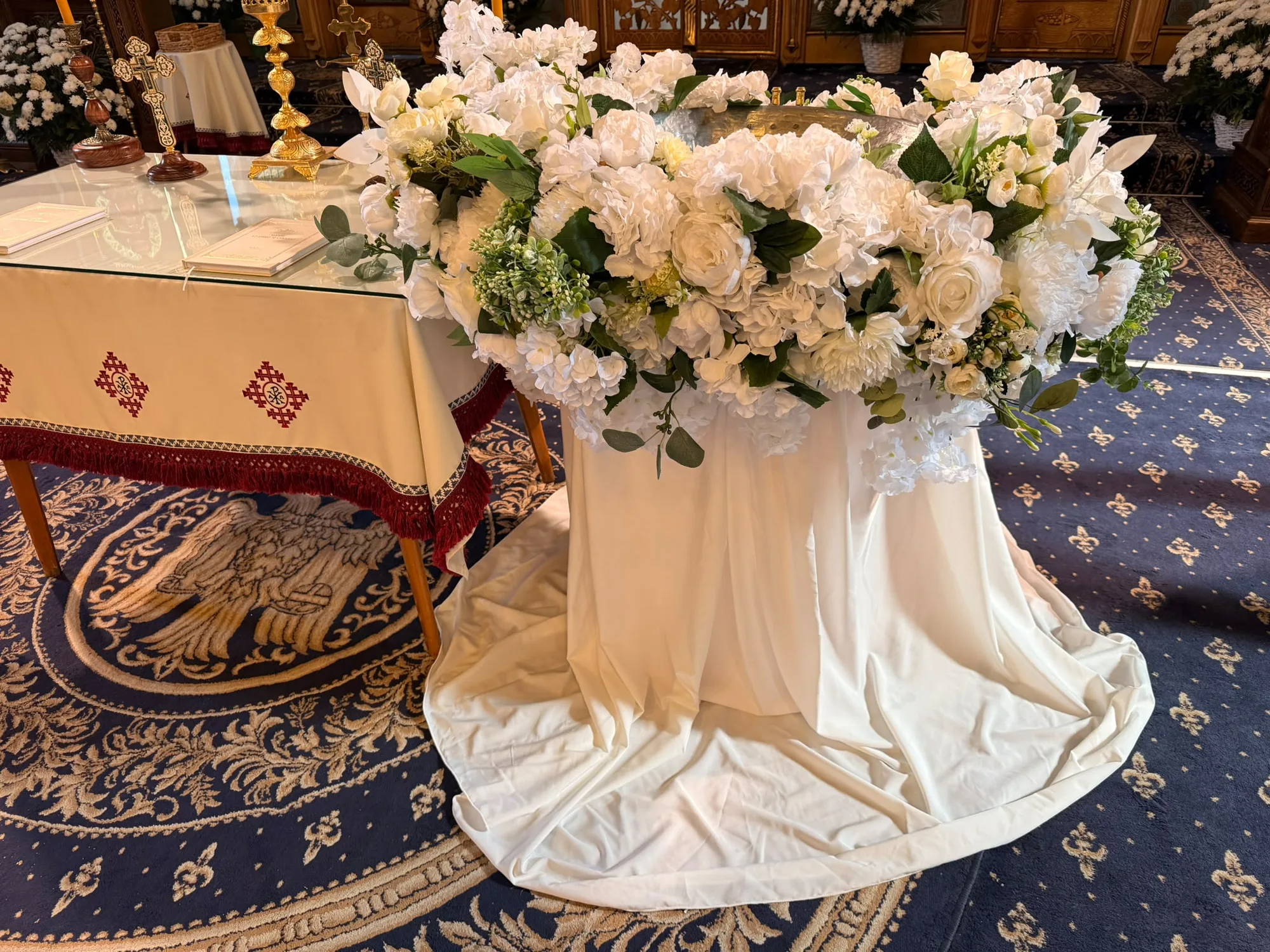Cristelniță decorată cu aranjament floral alb bogat și drapaj alb, în biserică