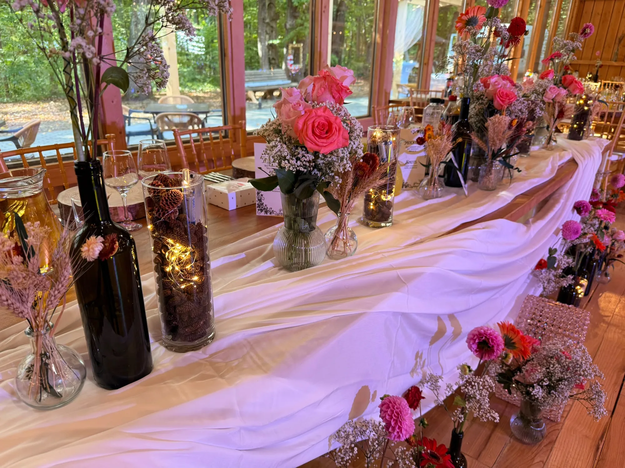 Masă lungă de nuntă decorată cu sticle de vin, flori roz, conuri de brad și luminițe