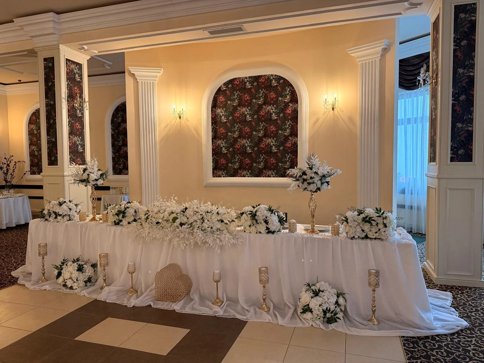 Prezidiu de nuntă lung cu flori albe și verdeață, în sală cu panouri florale pe pereți