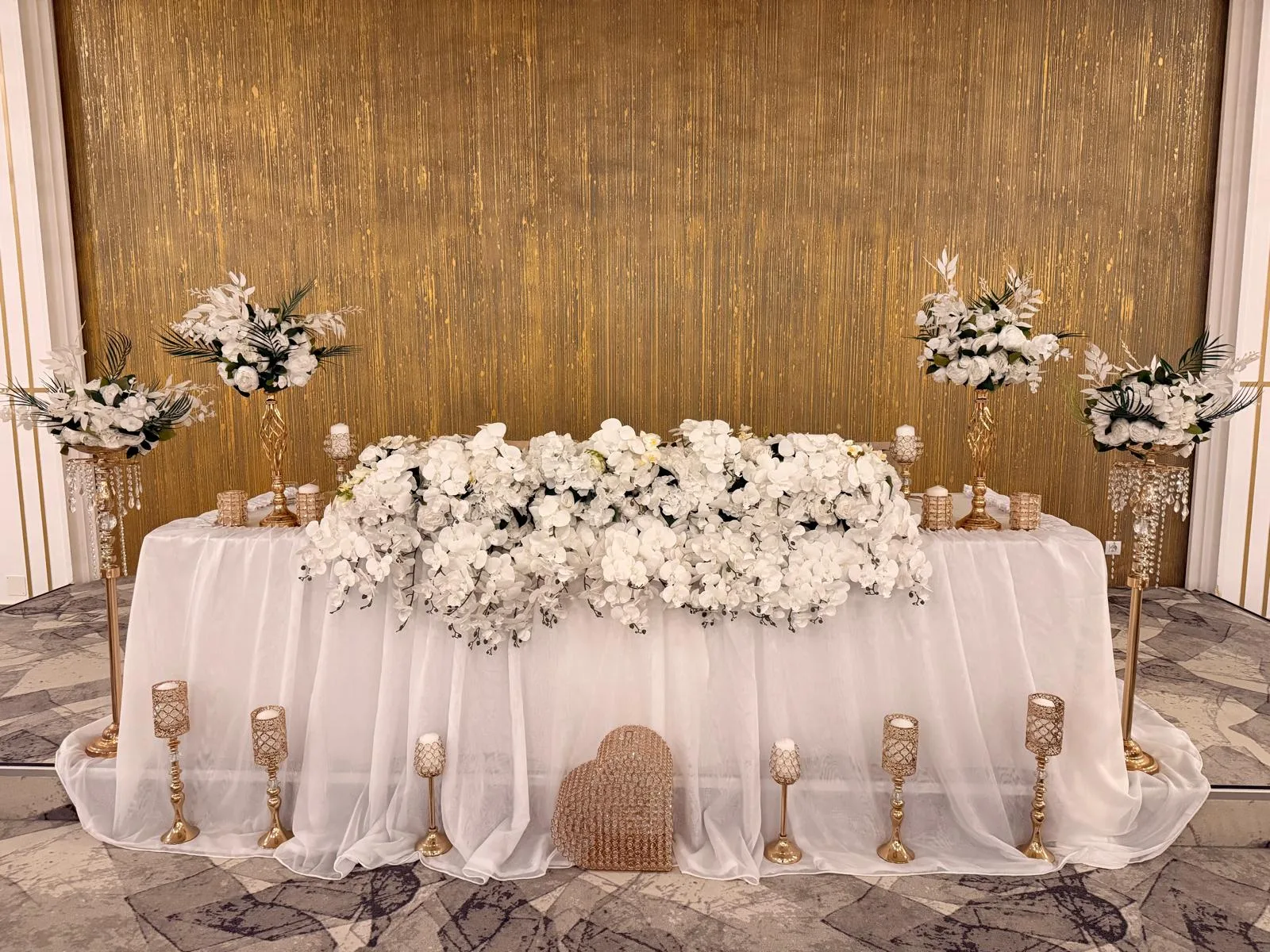 Prezidiu alb cu aranjamente florale pe stative aurii cu cristale si lumanari aurii, perete auriu striat