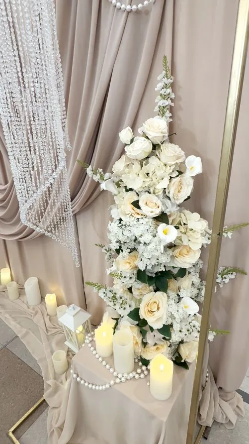 Aranjament floral alb-crem pe draperie bej, cu șiraguri de perle și lumânări