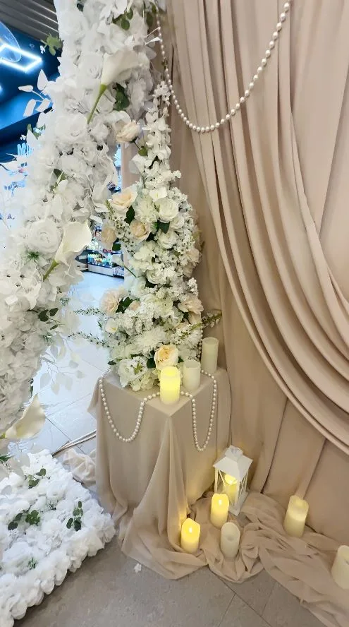 Perete cu draperie bej, flori albe agățate, șiraguri de perle și lumânări la bază