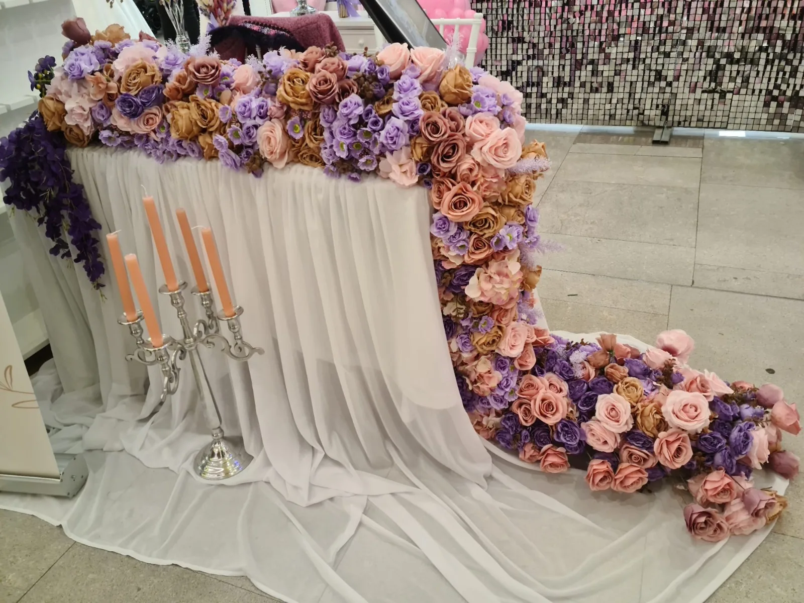 Masa cu trena lunga de flori mov-roz-auriu, draperie alba si candelabru cu lumanari