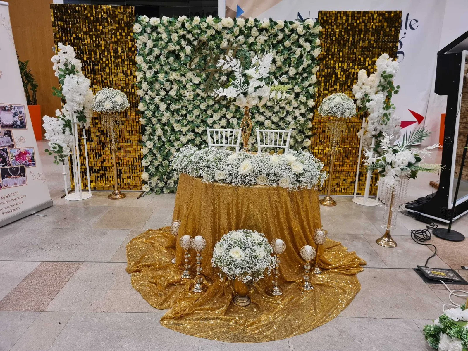Perete floral, masa aurie cu flori albe si candelabru auriu cu lumanari la targul de nunti