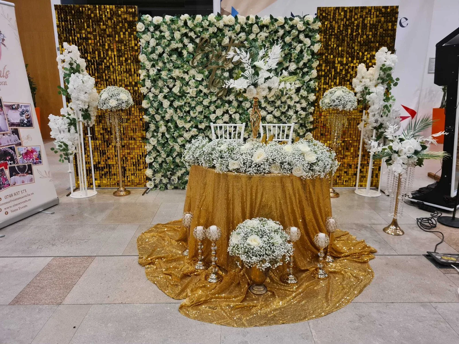 Detaliu perete floral verde-alb si masa aurie cu aranjament de flori albe la targul de nunti