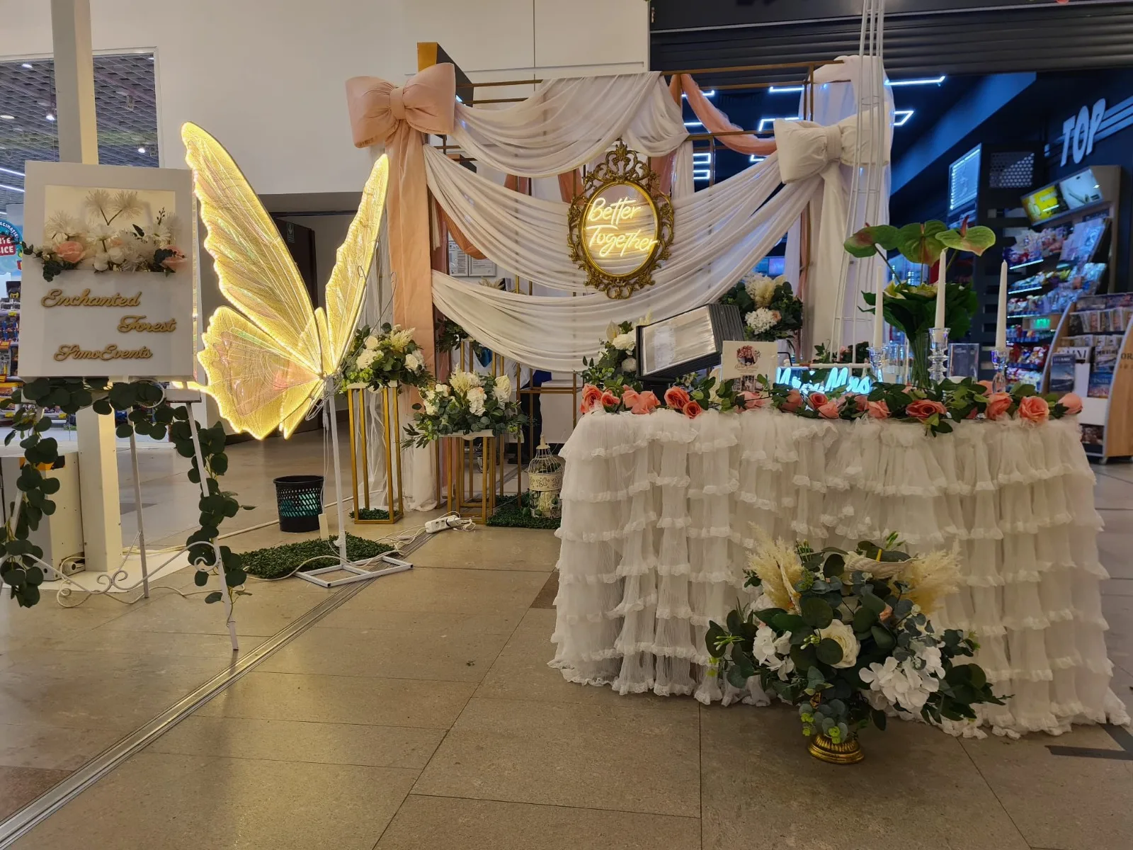 Panou Enchanted Forest, aripi de fluture si arcada cu fundita roz, langa masa cu flori albe si piersica